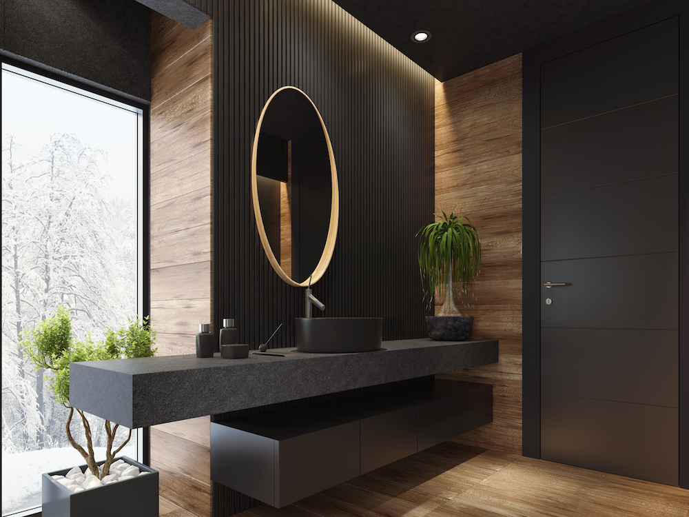 contemporary minimalist bathroom with black wooden walls and large grey matte and wood effect tiles. round mirror with wooden frame is on the wooden black wall. round grey stone washbasin and stainless steel basin tap is on top of the grey stone vanity unit with three drawers. freestanding black stone bathtub and floor mounted inox tap is on a platform with wood effect tiles. long floor tiles with wood effect. black ceiling with strip cove lighting and embedded spotlights. ***background is my istock image