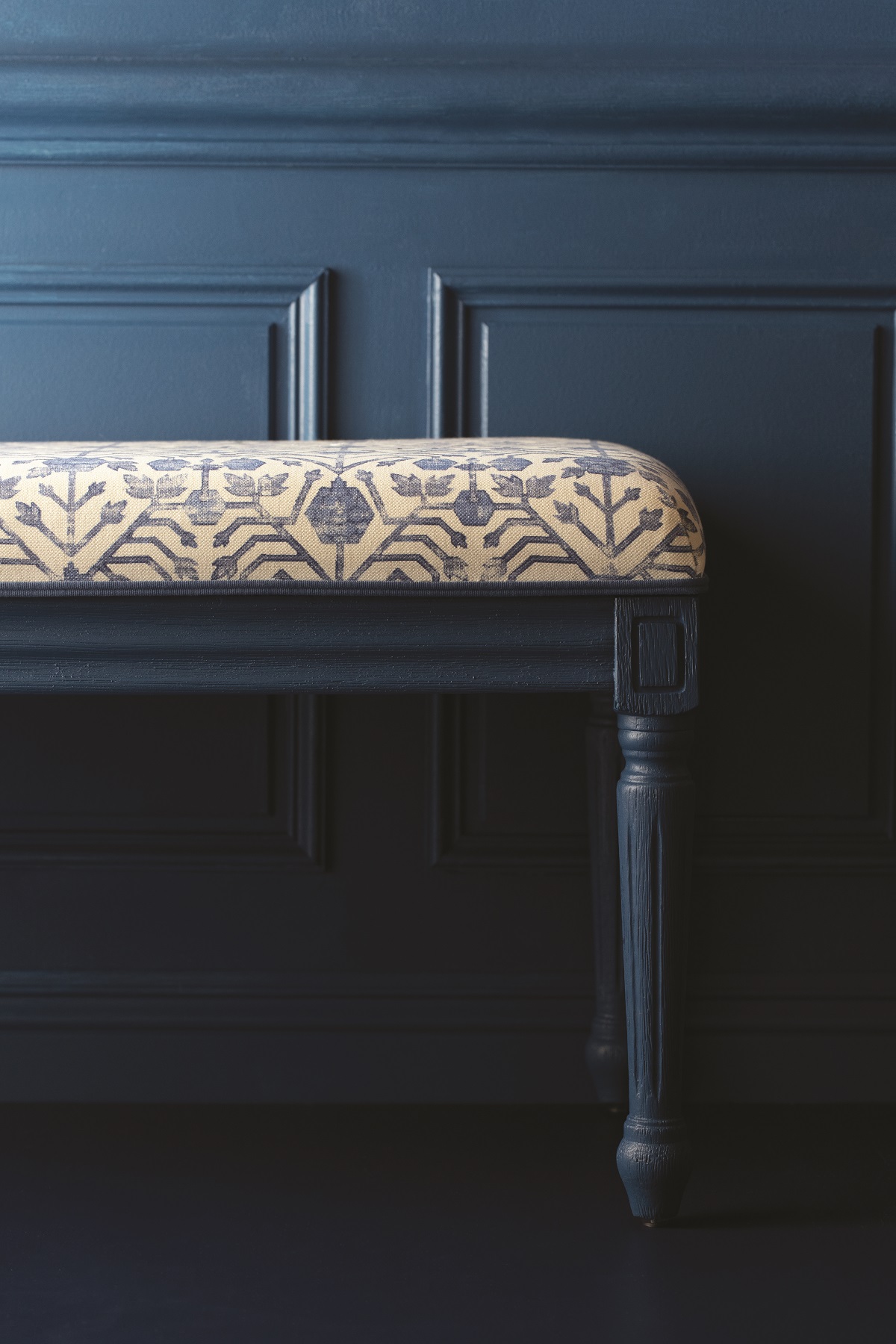 dark blue upholstered stool against adark blue wall with trim detail from Samuel & Sons