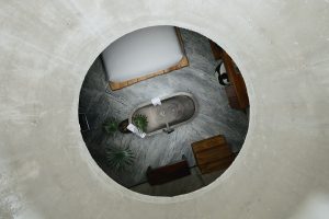 view through concrete structure and a round window down onto bathroom with central bath below the opening in Flavia Luxury Hotel by Rootstudio