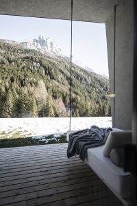 grey concrete and wooden surfaces with glass window looking out over mountains in front of a suspended hanging seat