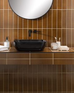 mustard coloured wall tiles from the Principle Collection on the vertical behind a round mirror and black handbasin