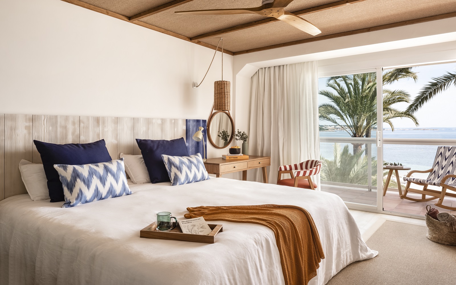 view across double bed with brown throw and blue cushions to windows and seaview in guestroom in Zel Mallorca