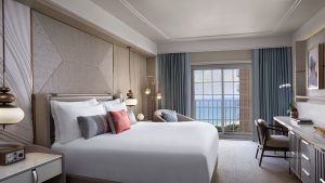 guestroom decorated neutral tones with blue curtains and a coral pink cushion with windows overlooking the sea