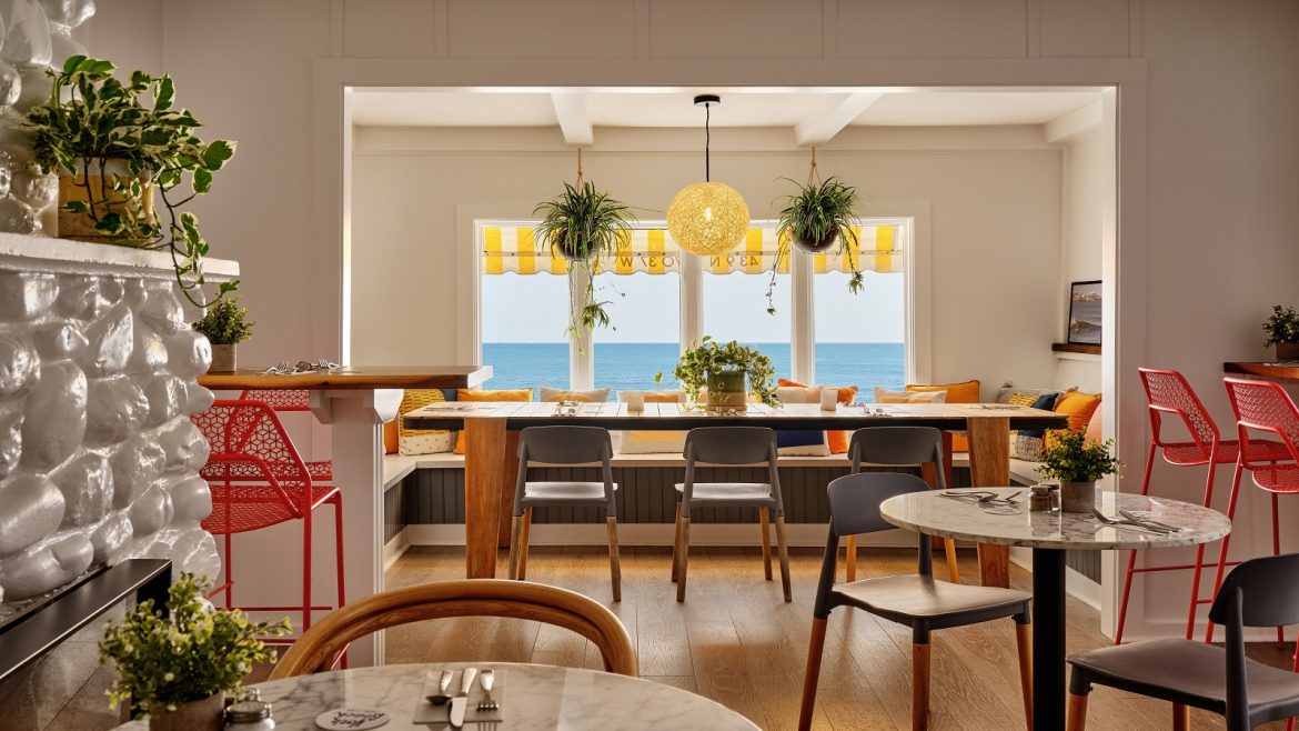 round tables with mid century chairs in front of a fireplace looking across to seating and a window out to sea at cafe in York Beach Surf Club