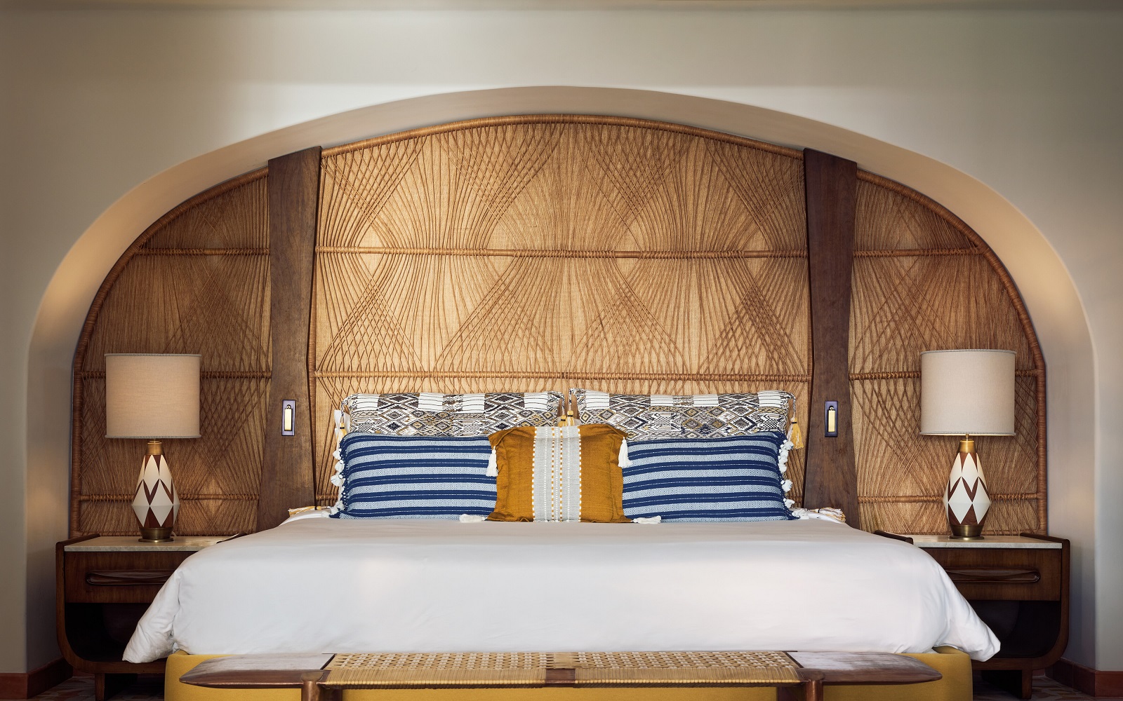 Maroma hotel bed set into arched alcove with a wicker woven headboard behind, blue striped pillows and ceramic bedside lamps