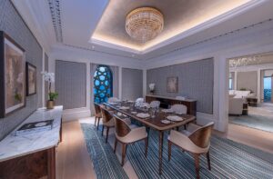 wooden dining room table and chairs under a chandelier and on top of blue textured carpet in guest suite