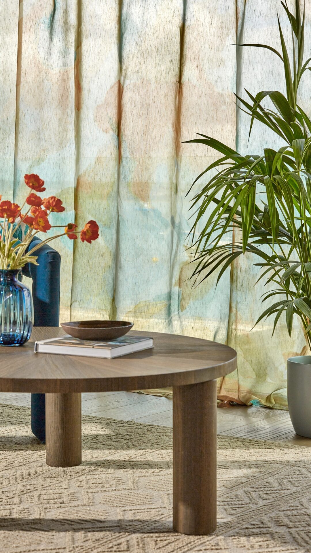 round wooden table with a vase of flowers in front of sheer curtains with a botanical print