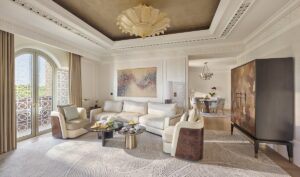 living room in hotel suite in shades of cream and gold with wooden accents and sand patterned carpet below recessed ceiling lighting