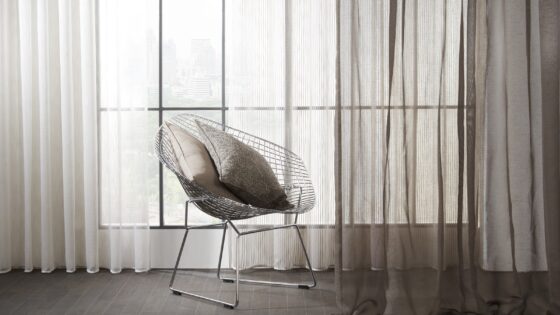 wire frame chair in front of a window with floor to ceiling voile curtains in mixed tones of grey