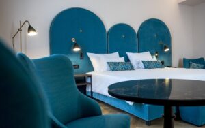 dramatic blue art deco curves of the upholstered blue headboard in the guestroom with blue furniture, white linen and contemporary lighting