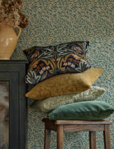 a pile of four cushions in patterned velvet on a wooden stool with Morris & Co wallpaper behind and a vase of flowers on a table on the side