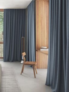 floor-to-ceiling grey curtains in bedroom with wooden chair and surfaces and stone tiled floor