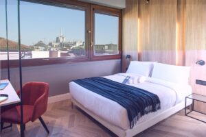 hotel guestroom with brown chair and grey throw on bed with view over Milan