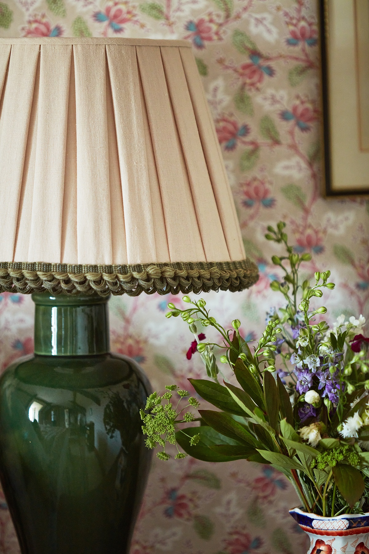 lampshade edged with green trim in front of floral wallpaper and vase of flowers