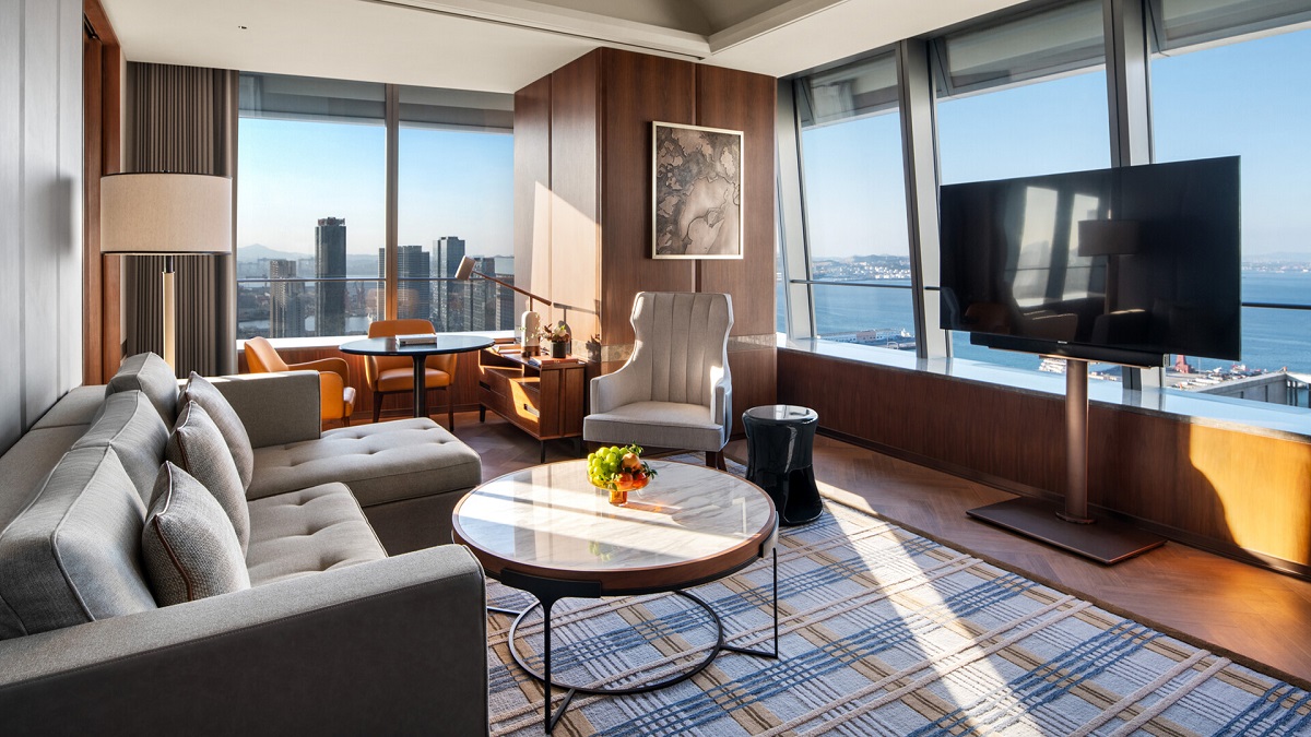 A cosy living area with a plump grey sofa, and marble-topped circular coffee table sitting atop a plaid rug with views across the city and ocean. 