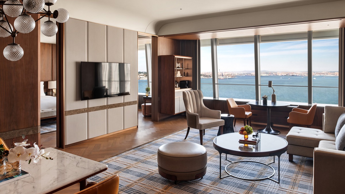 An executive room with warm, neutral coloured furniture, comfortable looking seating, and a large wall-hung television overlooks the ocean and city shoreline.