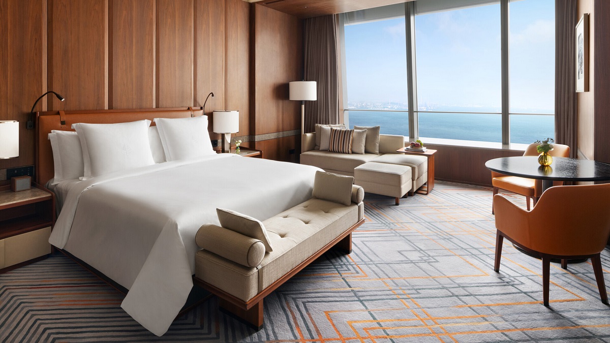 A guestroom where a king-sized bed with crispy white bedding takes centre stage with floor-to-ceiling wood panelling on the walls and window views of the ocean.