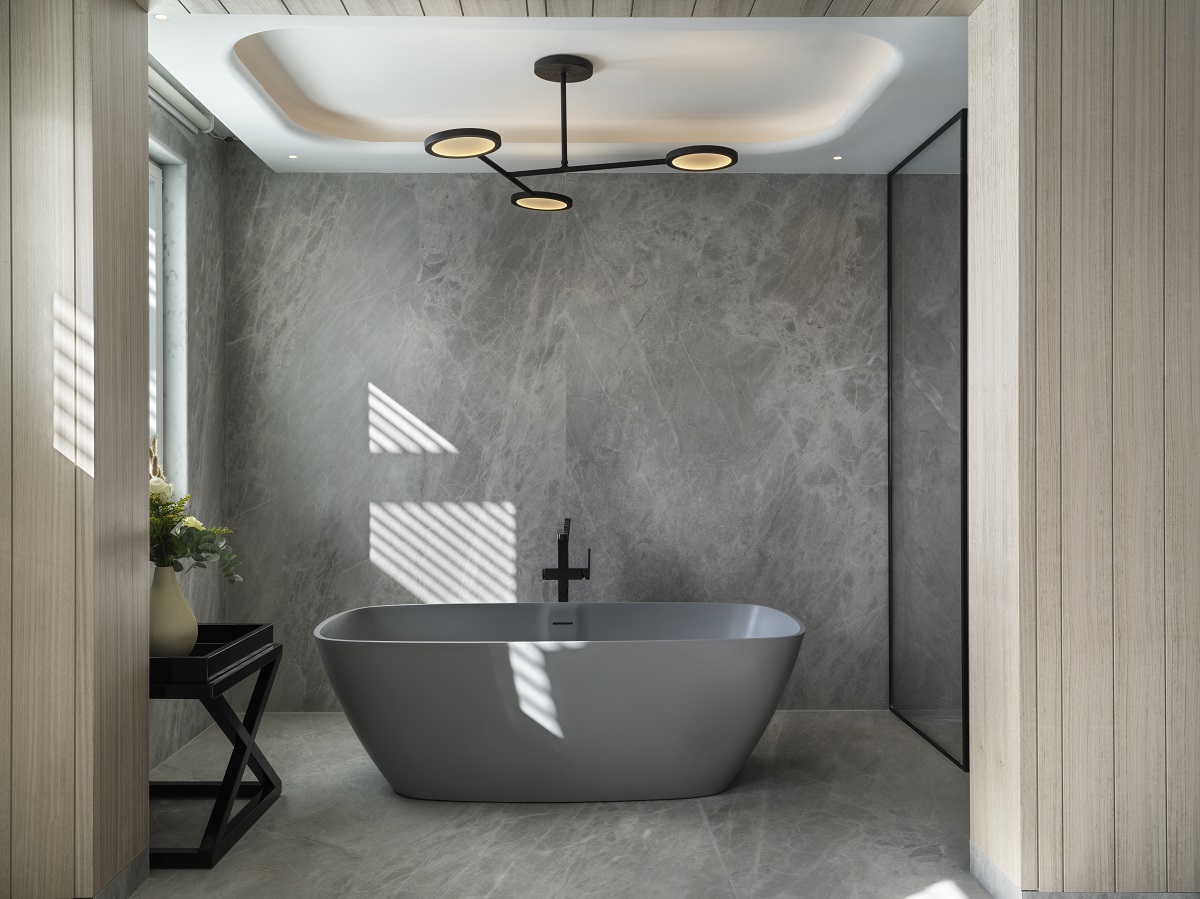 A chic bathroom setting with a largely grey palette where marbled walls and flooring set the stage for a large grey bathtub with feature lighting above. 