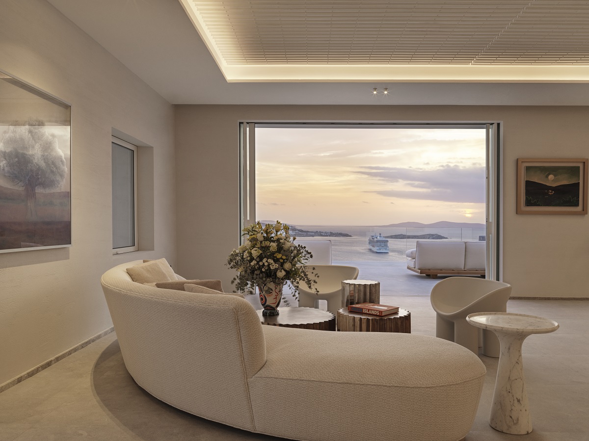 A soothing living room setting in a pale colour palette with curved sofa and sculpted furniture in front of open balcony doors framing views of the sea and neighbouring islands. 