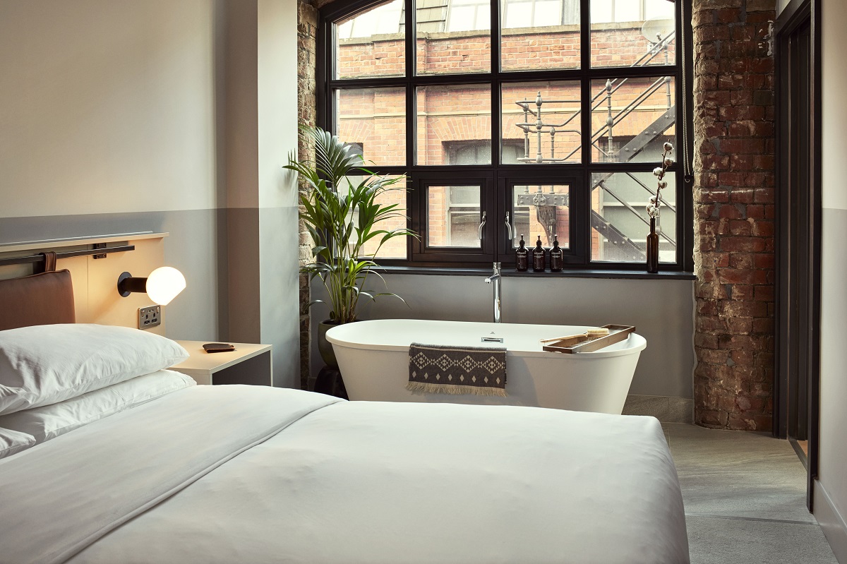 A guestroom at LEVEN Manchester showing industrial interior design, a cosy bed and standalone bathtub set under the room's window.