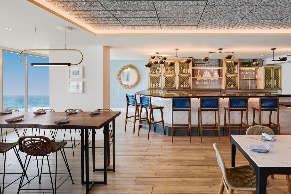 The lobby bar is a bright space with wooden flooring and a neutral colour palette of stone whites and cool blues. There is a bar at one side of the room and interesting angular lighting around the room, with sea views from floor-to-ceiling windows