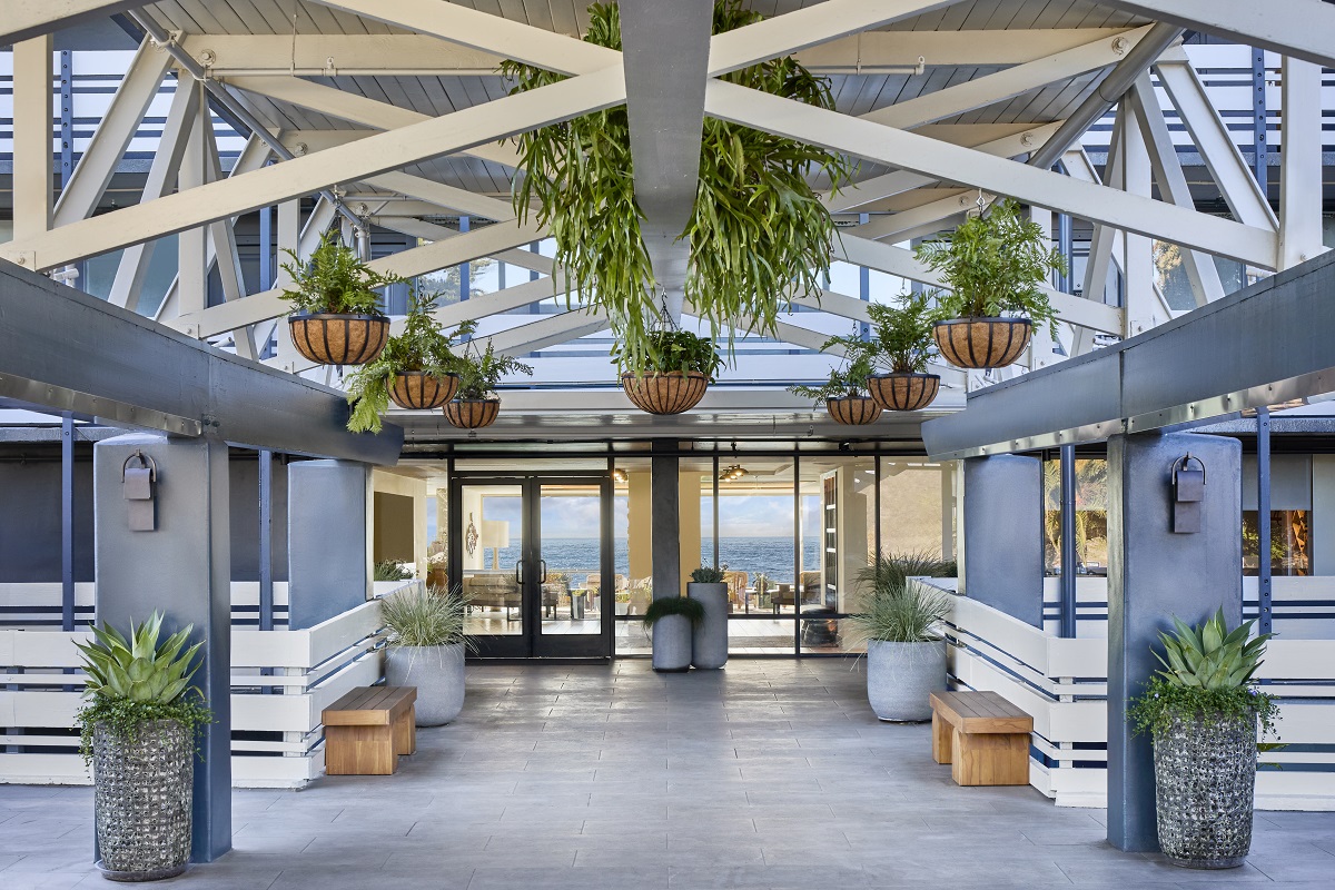 The entrance lobby consists of high ceilings and industrial exposed steel framework covered in foliage and hanging plants with views through to the other side of the building and the ocean beyond