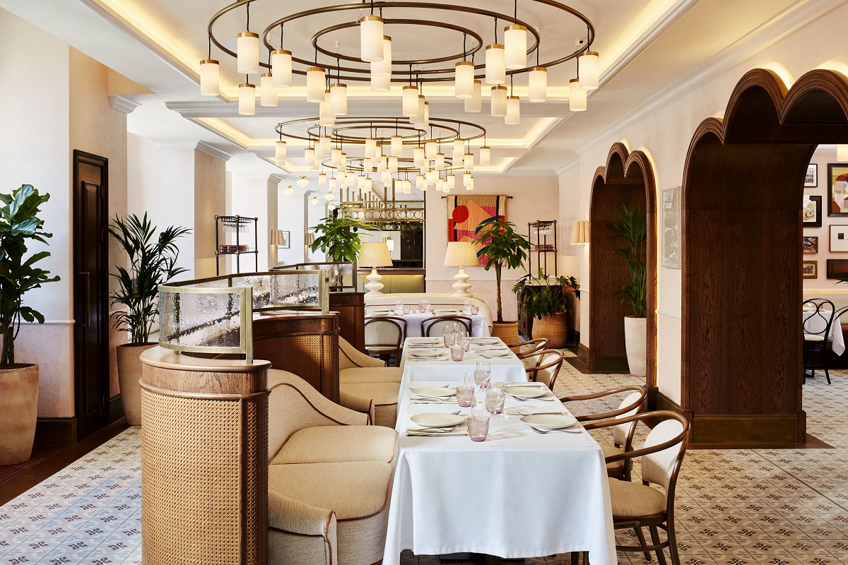 A chic dining area with a light and bright colour palette with neutral tones and natural materials. Large contemporary chandeliers light the tables below them and unusual wooden-clad curved openings separate different areas.