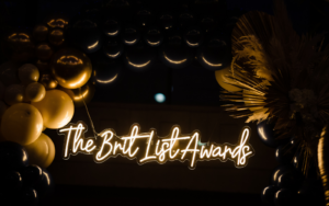 gold balloons and palm tree leaves surround a neon white and gold sign reading the brit list awards