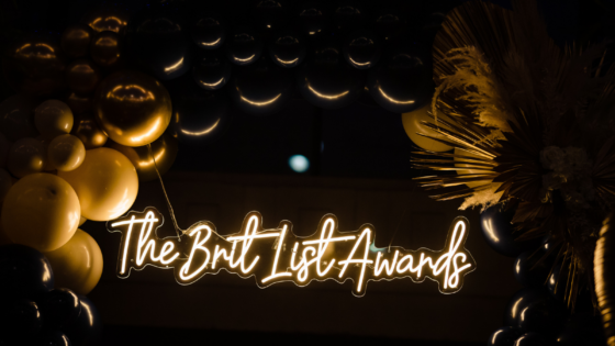 gold balloons and palm tree leaves surround a neon white and gold sign reading the brit list awards