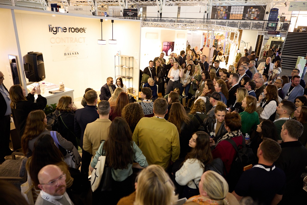 A large crowd gathers around the Ligne Roset and Balsan stand at HIX 2024 to listen to a talk entitled 'Designing the Brand' hosted by Sophie Harper with guest speakers Henry Reeve, Arun Rana, Grace Kelly, and Justin Meath-Baker. 