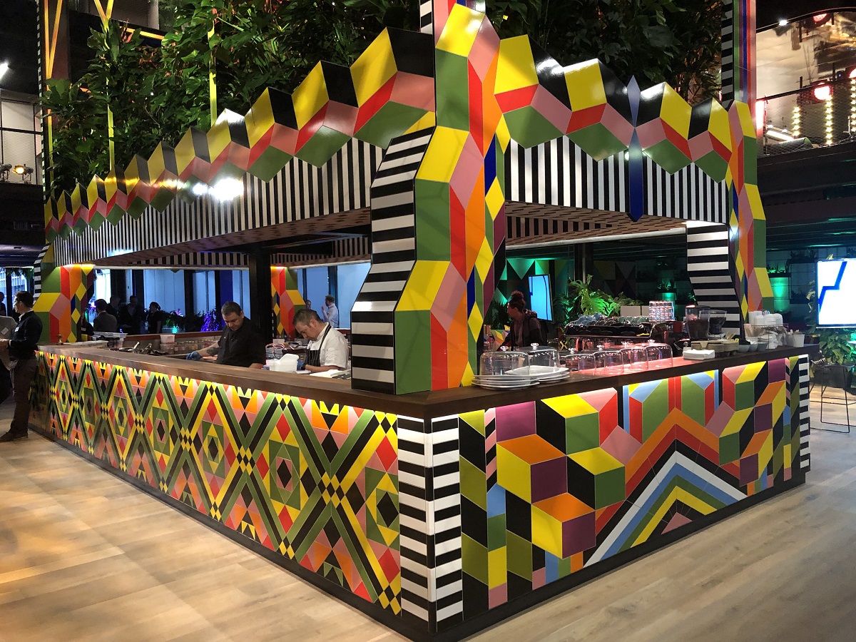 A pop-up serving space for food and drinks within an exhibition space has been elaborately decorated with colourful tiles in lots of colours and patterns, with thick foliage sprouting out of the roof.