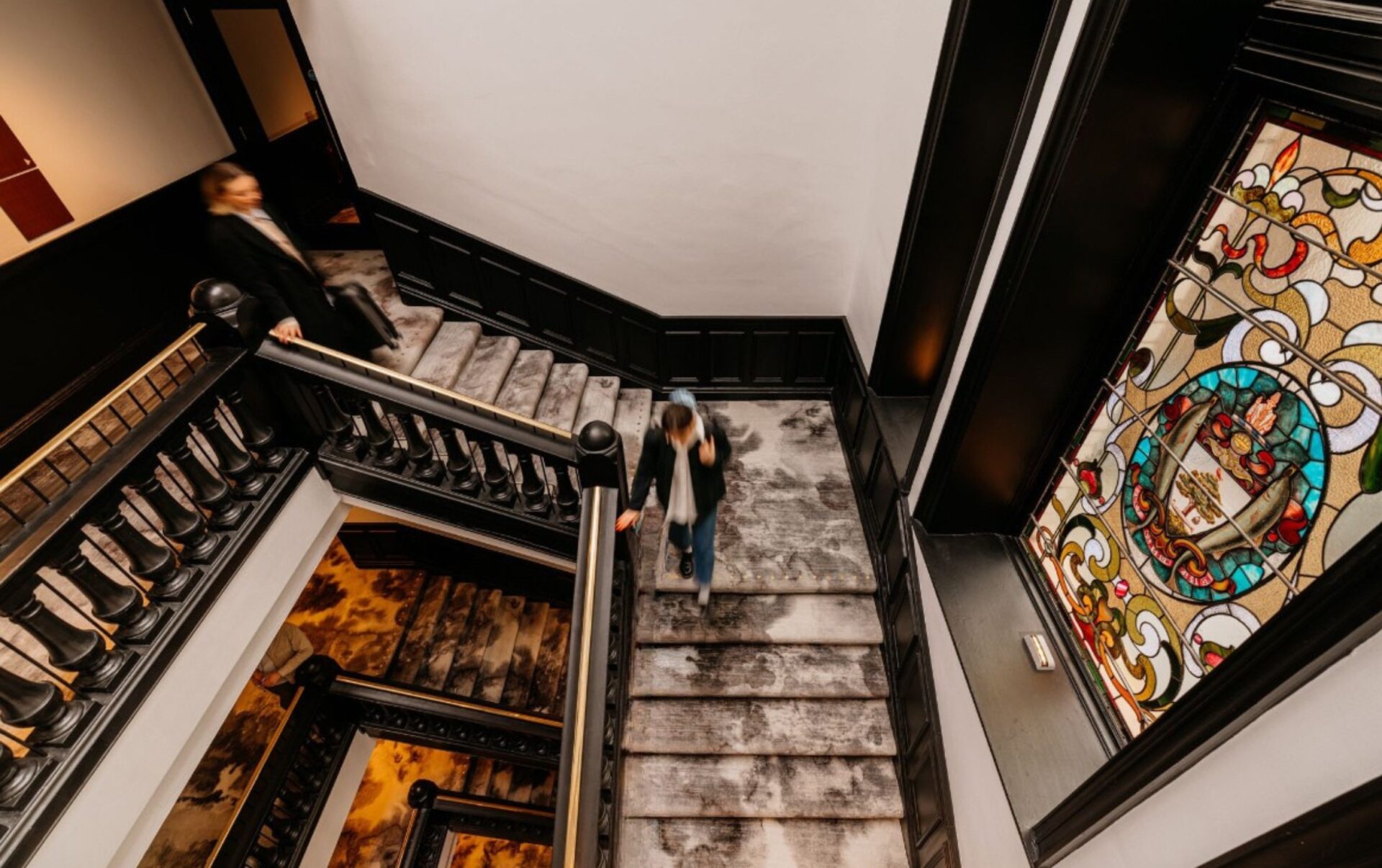 Birds eye view of square staircase, from several floors above. Grey patterned carpet lines the stairs. There is a stained glass window to the right of the staircase. 