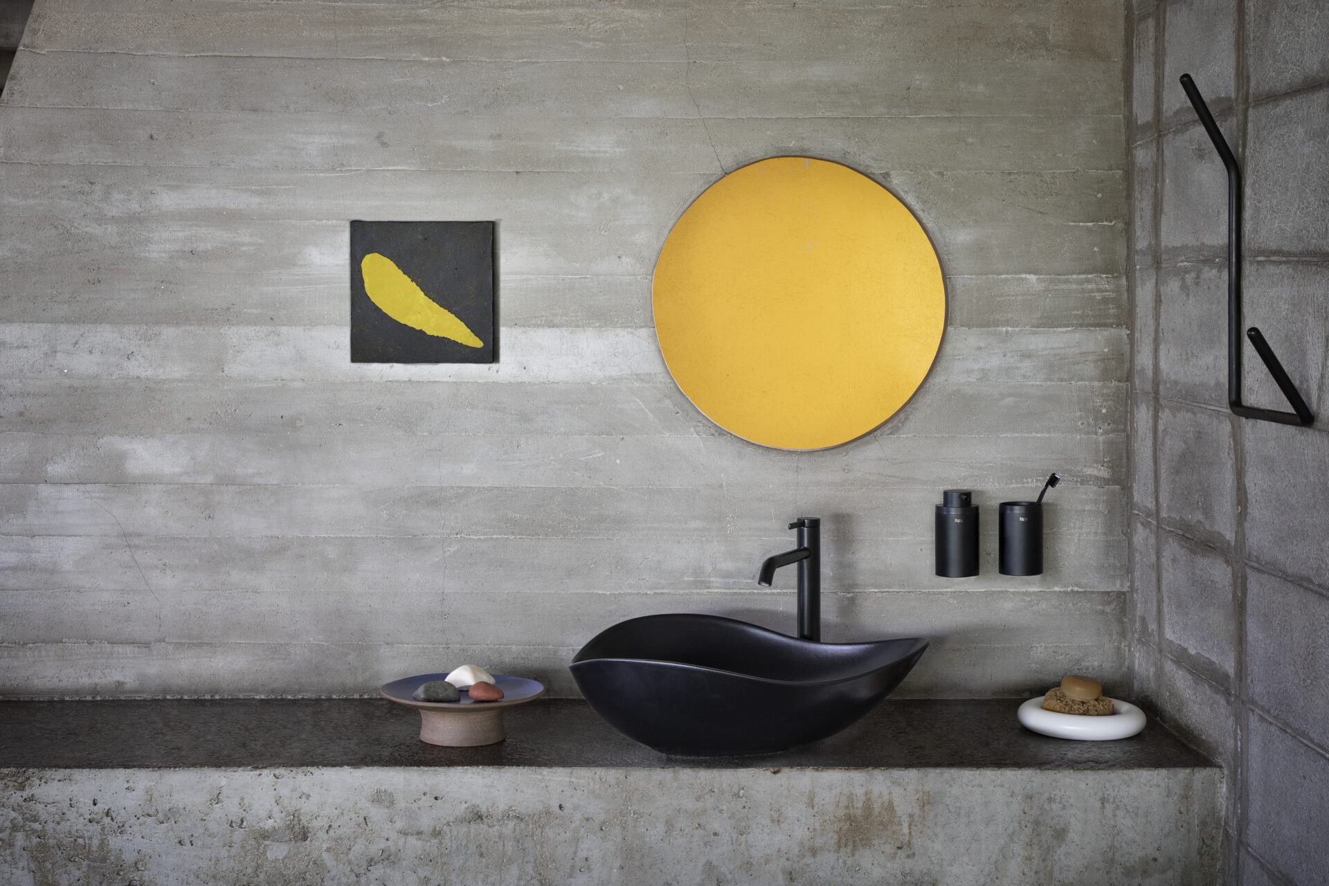 A modern minimalist bathroom with a black sculptural sink, a black faucet, and concrete walls. A large round yellow mirror hangs above the sink, accompanied by a small black wall-mounted soap dispenser and toothbrush holder. To the left, a black square artwork with a yellow abstract shape is mounted on the wall. The countertop features a ceramic dish with smooth stones and a white plate holding a natural sponge and soap. The space has a raw, industrial aesthetic with a neutral color palette accented by bold black and yellow elements.