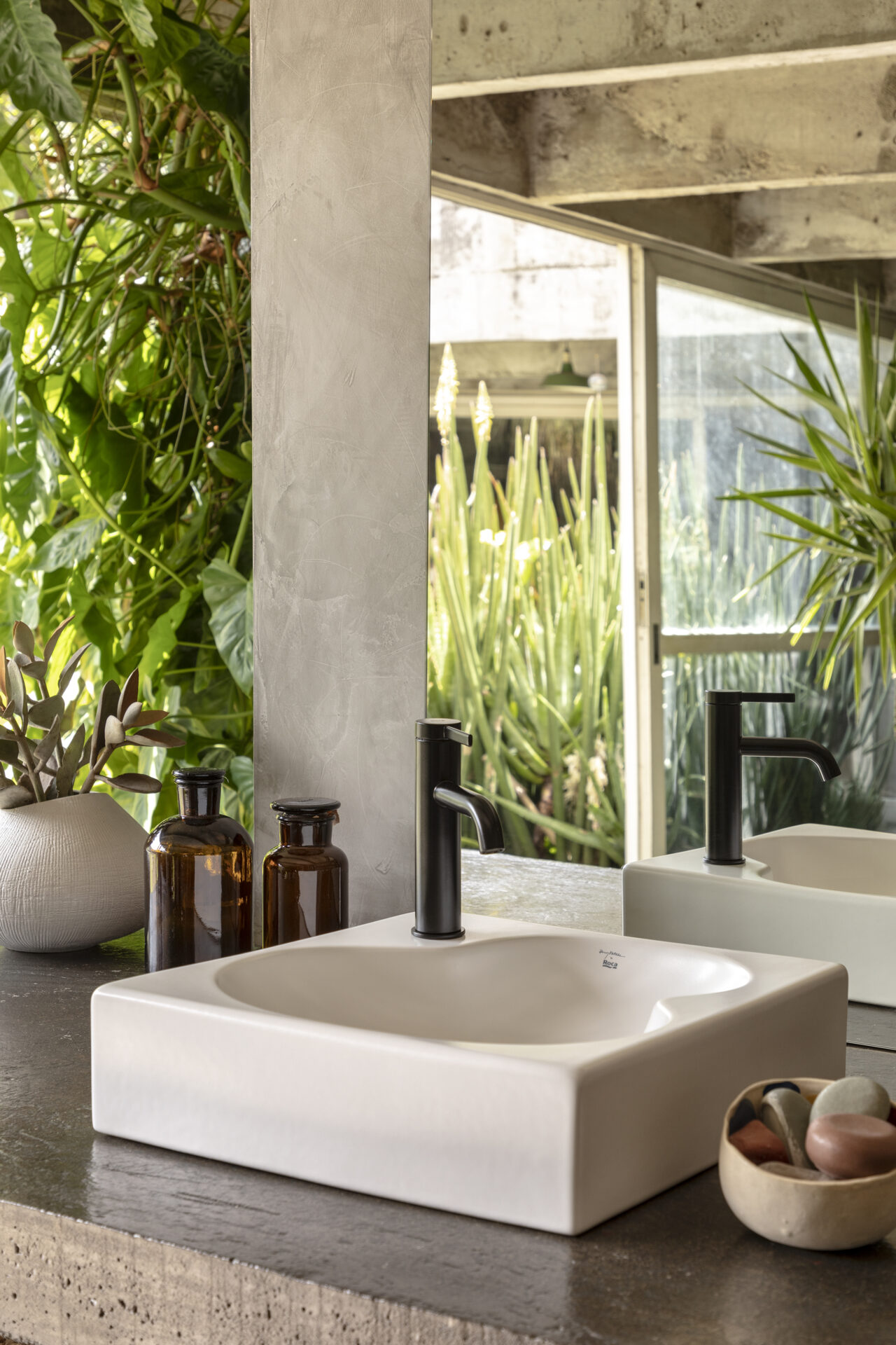 A modern bathroom sink area with a white square-shaped basin and a sleek black faucet mounted on a textured concrete countertop. The space is decorated with amber glass bottles, a white ceramic vase with plants, and a small bowl of smooth stones. A large mirror reflects lush greenery from the outdoor garden, creating a serene and natural ambiance. The design combines industrial and organic elements with a neutral color palette and natural textures.