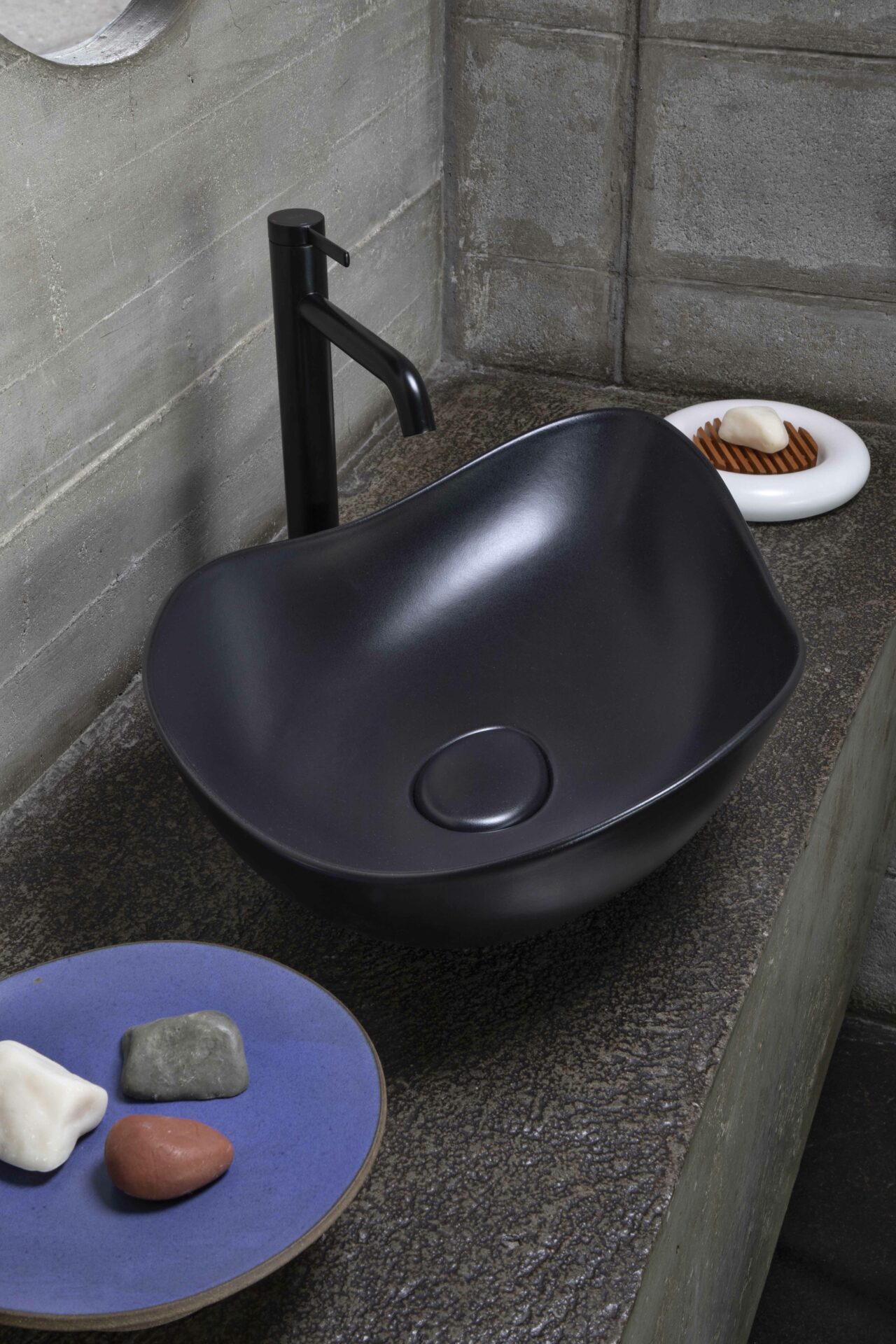 A contemporary bathroom setup featuring a black sculptural vessel sink with a matte finish and a matching black faucet. The dark textured countertop contrasts with the raw concrete walls. A deep blue ceramic plate with multicolored stones and a white soap dish with a wooden insert add subtle decorative elements. The minimalist design and organic shapes create a sophisticated, spa-like atmosphere.