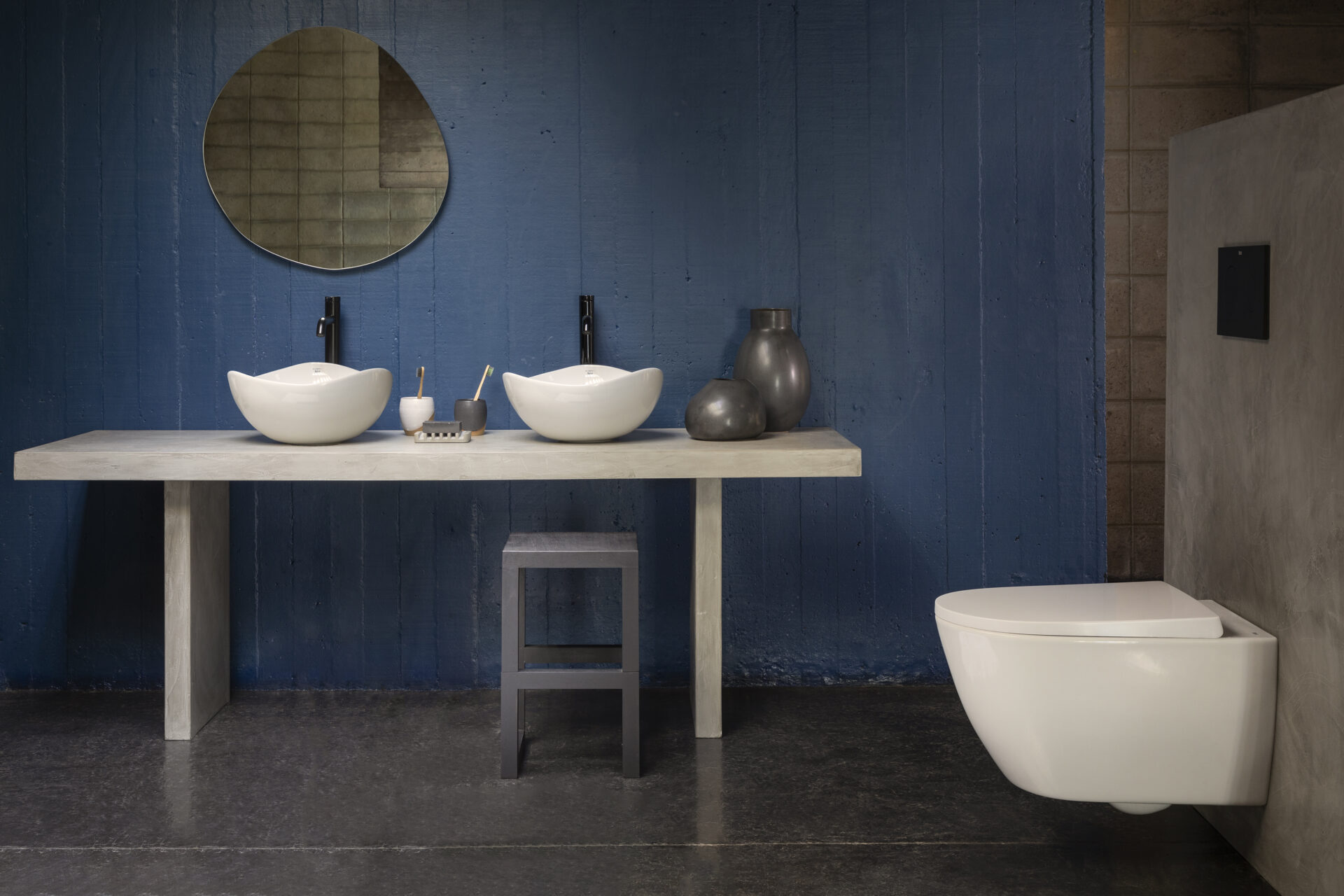 A contemporary bathroom set up with blue walls and an organically shaped mirror above dual white sculptural sinks. A white floating toilet sits against a stone wall. 