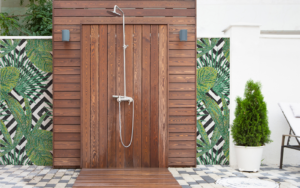 Outdoor shower area, with shower mounted on slatted wooden panel, surrounded by mosaic decorations which shows a green leaf pattern overlaid on a black and white geometric pattern