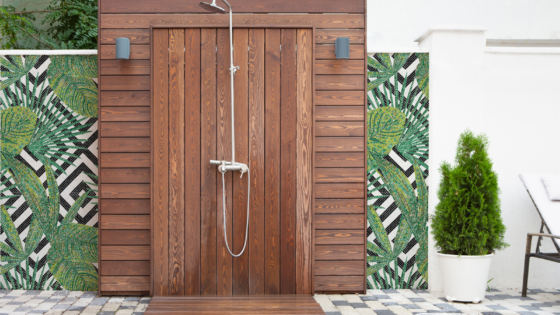 Outdoor shower area, with shower mounted on slatted wooden panel, surrounded by mosaic decorations which shows a green leaf pattern overlaid on a black and white geometric pattern