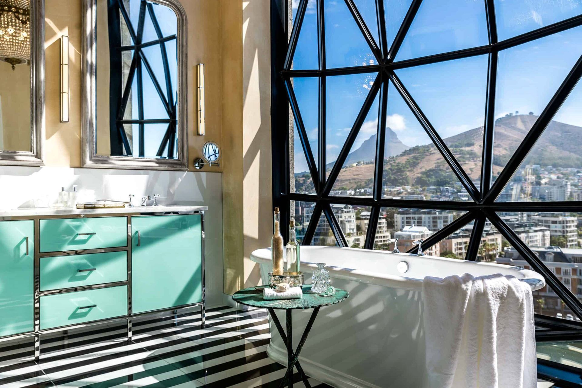 A luxurious bathroom with a floor-to-ceiling geometric glass window offering a stunning view of Cape Town, including the cityscape and surrounding mountains. The bathroom features a freestanding white bathtub with a plush white towel draped over its edge, a sleek vanity with mint-green cabinets and a marble countertop, and a small round table holding decorative bottles and a towel. The black-and-white striped floor adds a bold contrast to the elegant design, and natural light streams in, enhancing the airy ambiance.