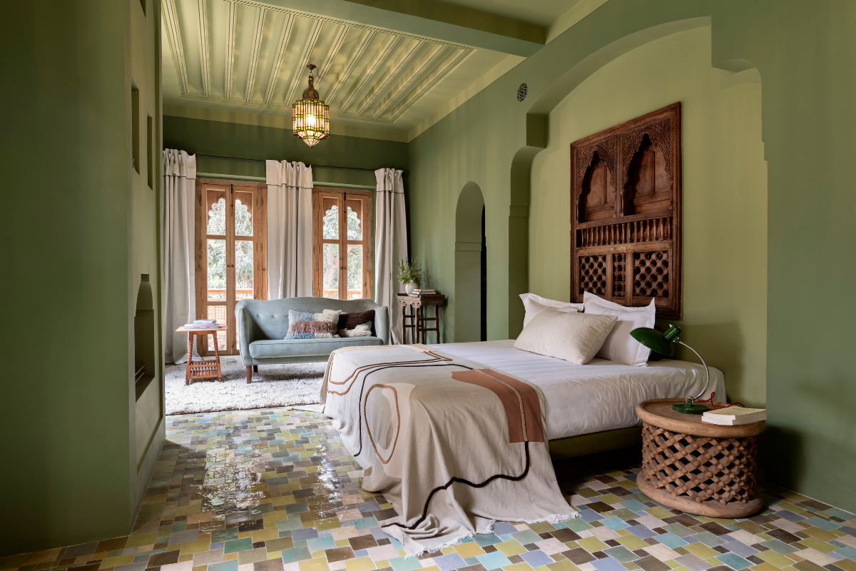 Green bedroom with cream, blue and brown glazed tile floor. Bed in foreground faces built in fireplace while sofa sits infront of floor to ceiling windows at far end of room