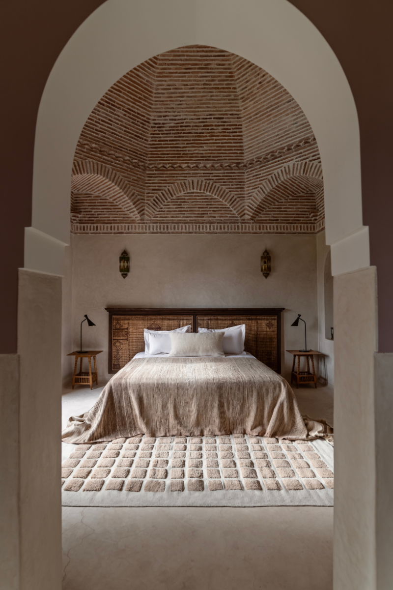 Shot through arched doorway into neutral toned bedroom with domed ceiling with rattan-look walls. Bed sits central in room