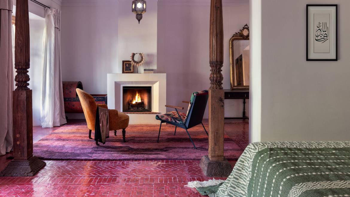 Guestroom at the Jnane Rumi in Marrakech, with pink tiled floor before wooden framed entrance to seating area with two armchairs infront of open fire. Floor turns to magenta and lilac woven rug
