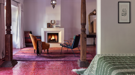 Guestroom at the Jnane Rumi in Marrakech, with pink tiled floor before wooden framed entrance to seating area with two armchairs infront of open fire. Floor turns to magenta and lilac woven rug