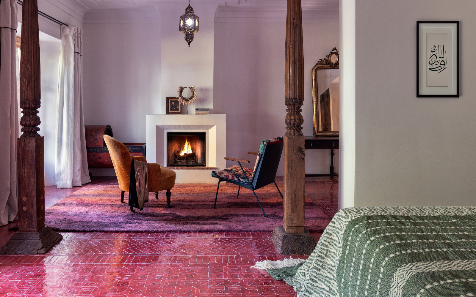 Guestroom at the Jnane Rumi in Marrakech, with pink tiled floor before wooden framed entrance to seating area with two armchairs infront of open fire. Floor turns to magenta and lilac woven rug