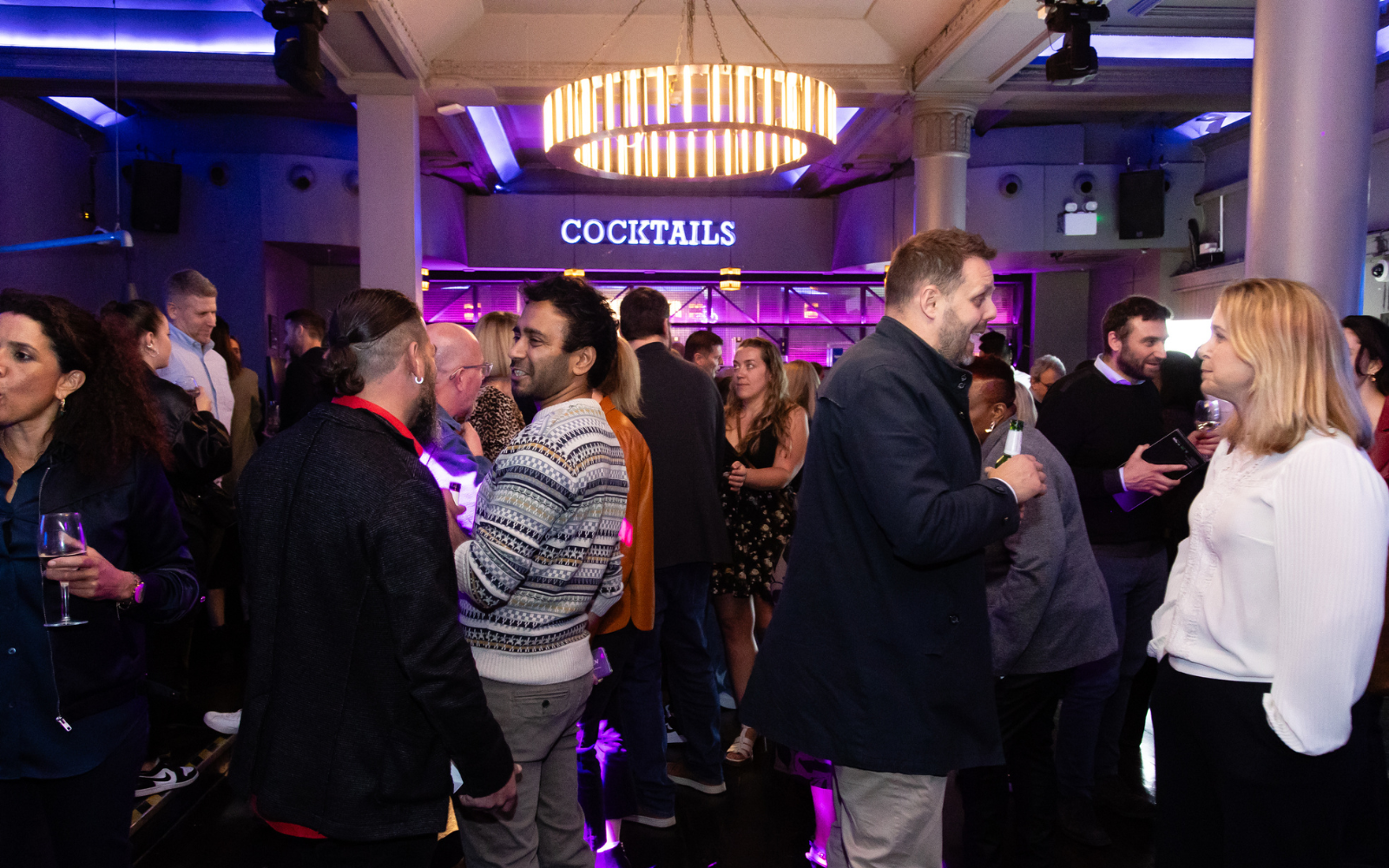 Group of MEET UP London attendees stood infront of neon lit cocktail sign and bar