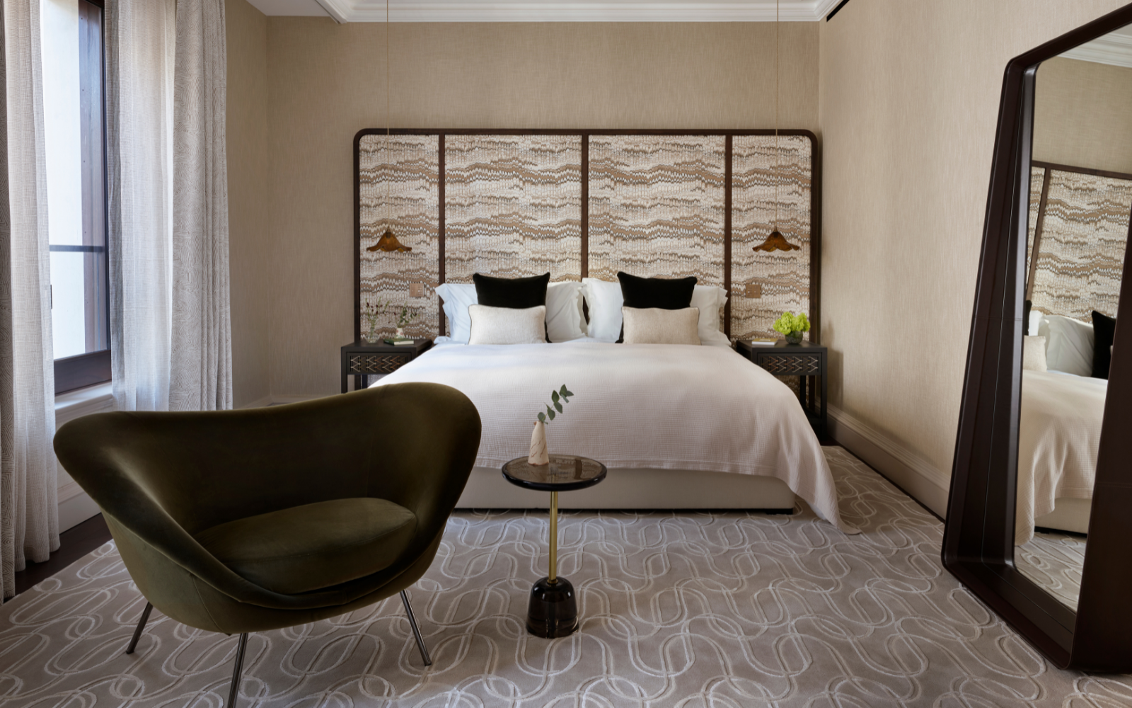 Neutral-toned bedroom, mid-century modernism in design, in The OWO Residences by Raffles. Textured headboard frames the bed, while a tall mirror leans against the wall to the right and a organic shaped armchair sits to the left of the bed. By StudioMorey