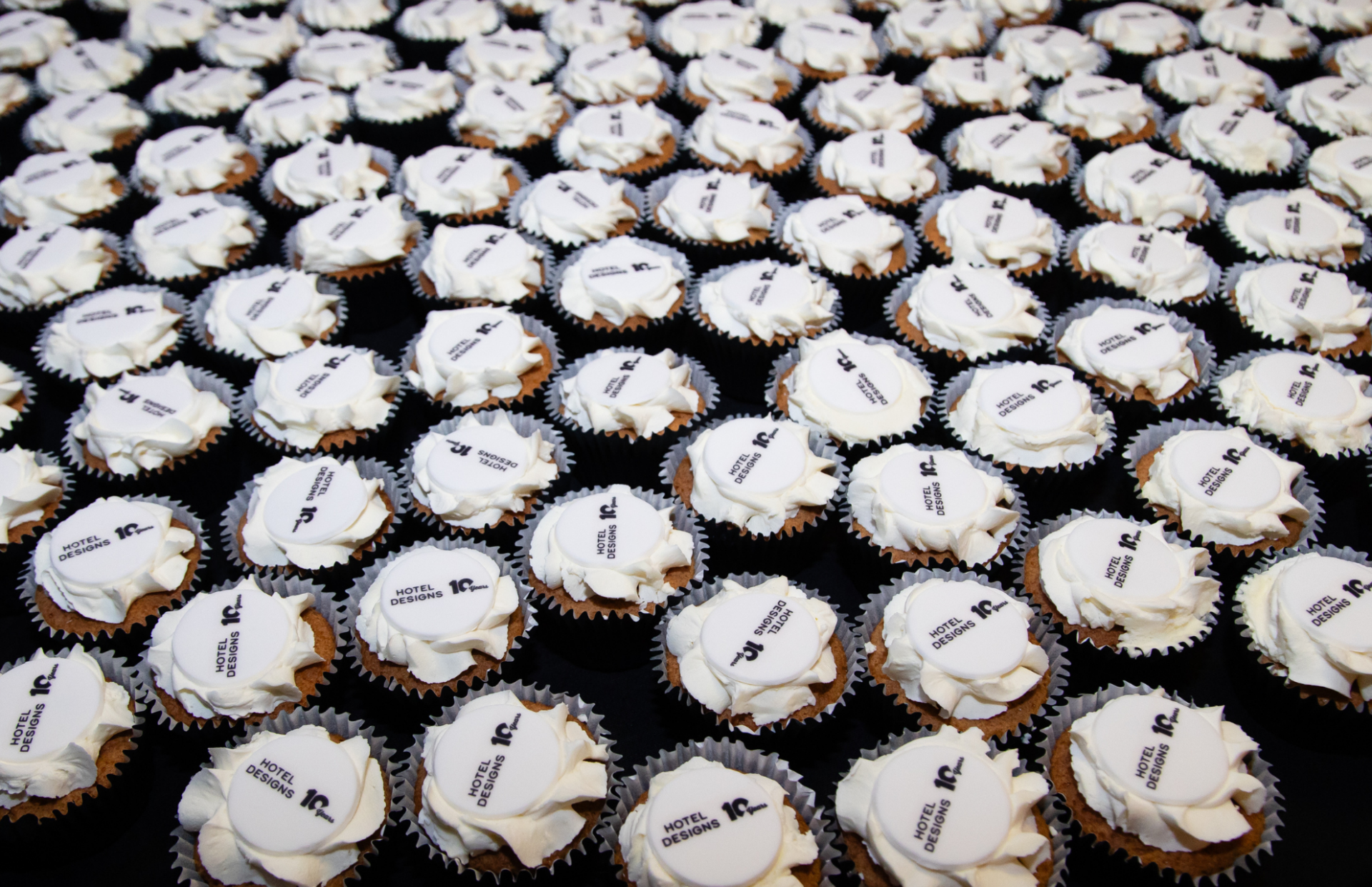 Table full of white cupcakes with Hotel Designs 10 Years logo on each cake