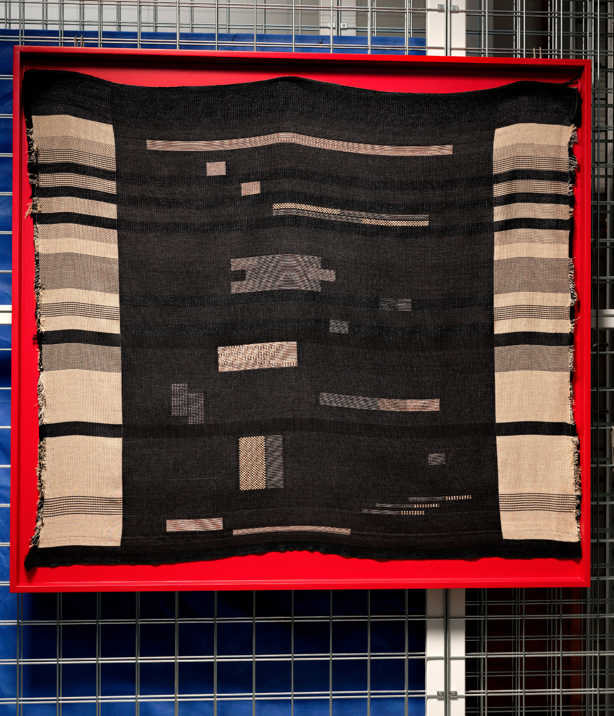 Greyscale woven fabric on red backdrop, hanging from metal fencing