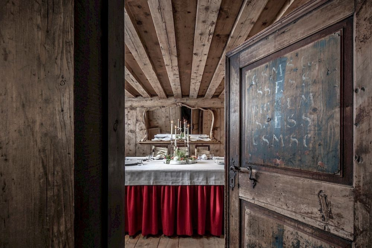 Dining area in Hotel La Perla_Corvara, Italy, wooden ceiling and walls surround a square table with red fabric trim and white table cloth. Chairs with ribbon woven through backrest sit on three sides and candelabra in the middle, from The Leading Hotels of the World new book