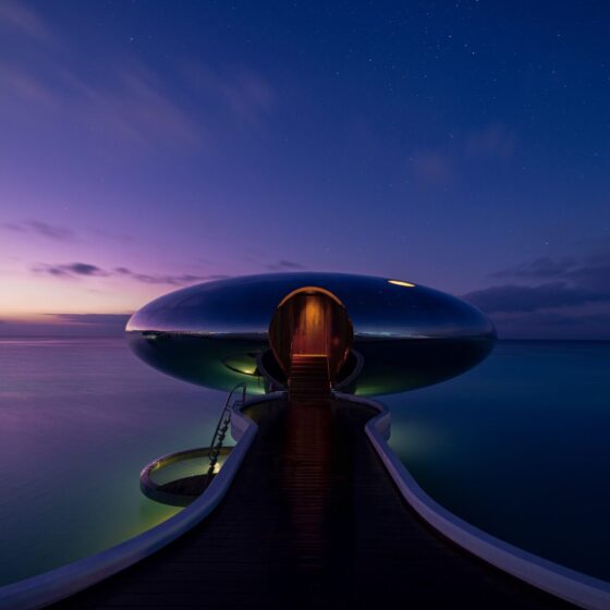 Silver pod hotel room shot at night with dark inky purple-blue skyline and sea. Orange light beams down from pod front door